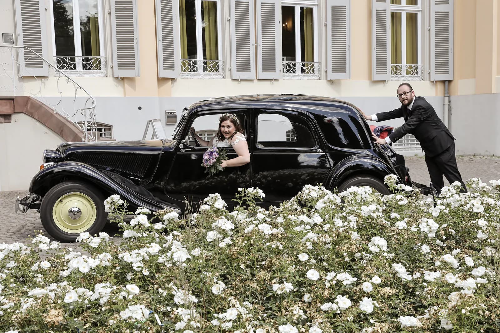 Braut mit Blumenstrauß lehnt aus einem schwarzen Oldtimer, der Bräutigam schiebt von hinten.