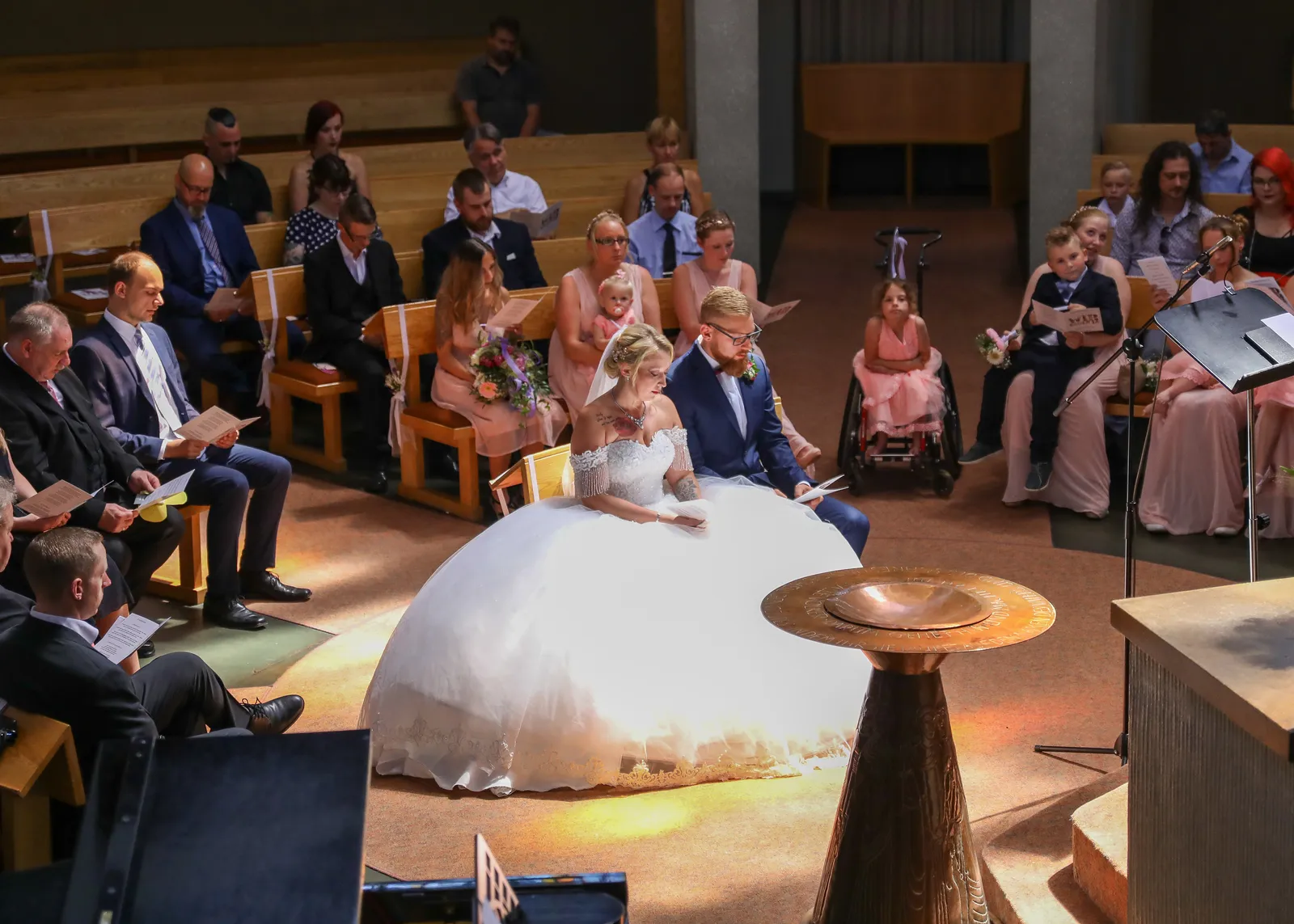 Brautpaar sitzt in einer Kirche vor den Gästen, die Liedblätter in den Händen halten.