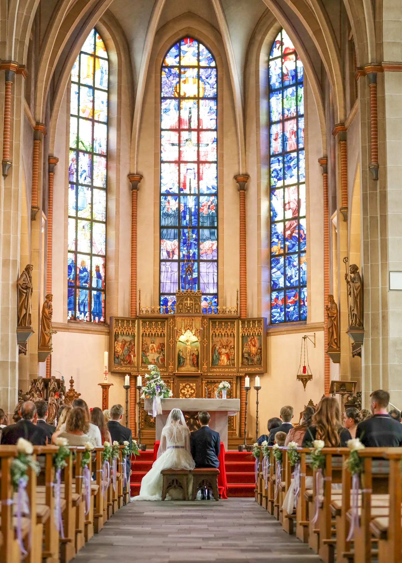 Brautpaar kniet vor dem Altar einer Kirche, dahinter hohe bunte Glasfenster, Gäste in den Bänken.