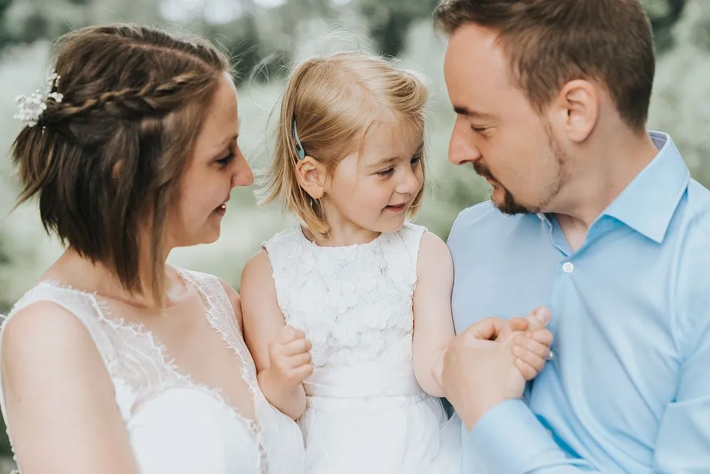 Brautpaar im Freien, zwischen ihnen ein kleines Mädchen im weißen Kleid.