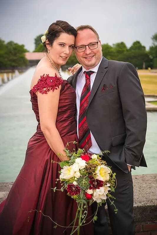 Frau in dunkelrotem Kleid mit Blumenstrauß neben Mann im grauen Anzug vor einem langen Wasserbecken.