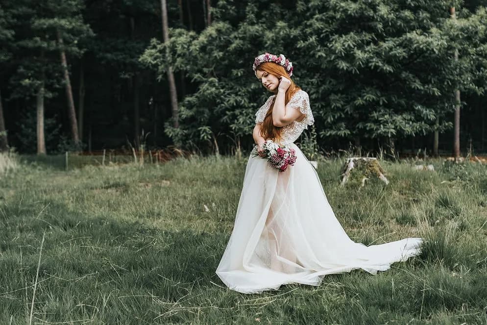 Frau in weißem Tüllkleid mit Blumenkranz und Strauß auf einer Wiese vor Nadelwald.