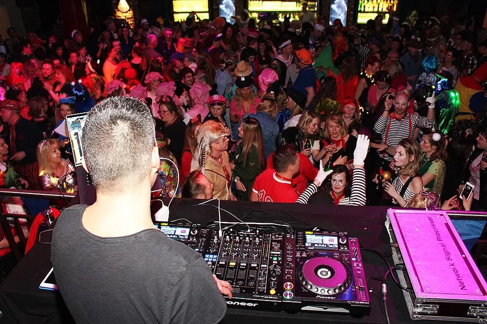 DJ von hinten am Pult, davor dicht gedrängtes Publikum in bunten Kostümen.