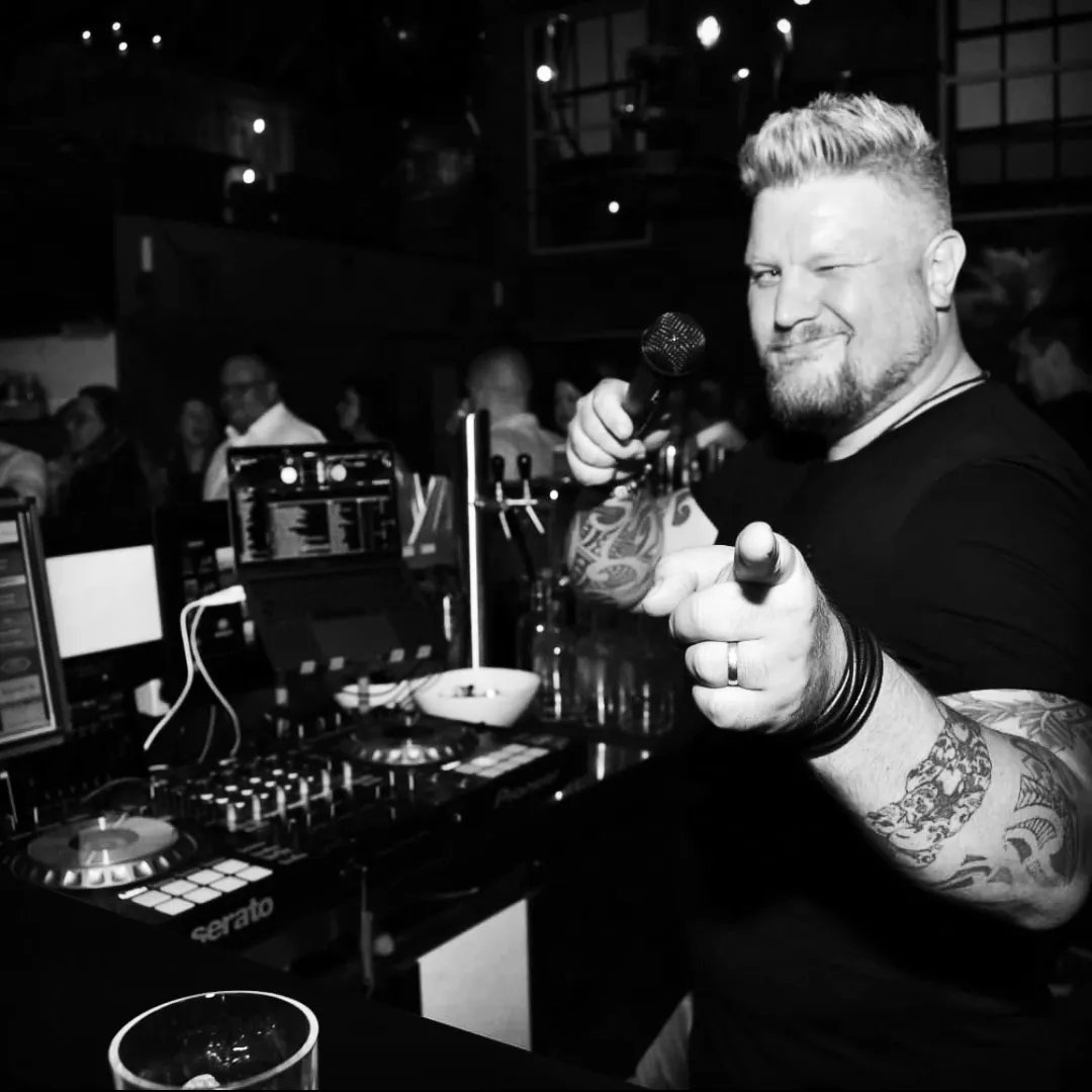 DJ Smatzo hält am Pult ein Mikrofon in die Kamera, tätowierte Arme, Schwarz-Weiß-Aufnahme.