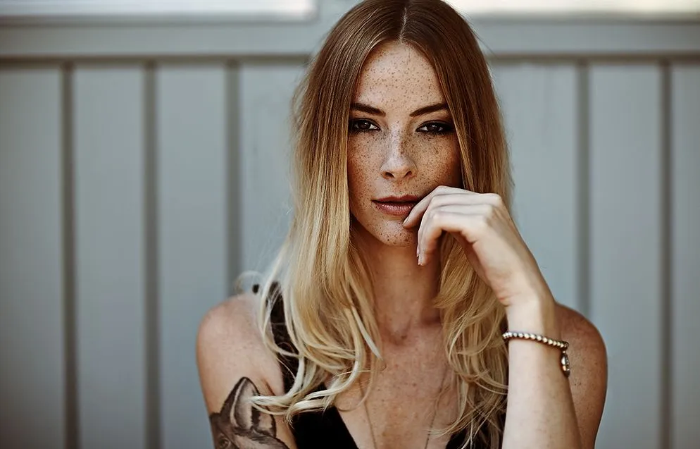 Porträt einer blonden Frau mit Sommersprossen, Hand am Kinn, vor heller Holzwand.