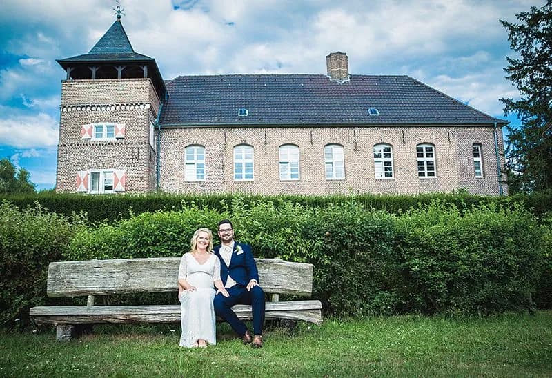 Paar in Brautkleid und Anzug auf einer Holzbank vor einer Hecke, dahinter ein Backsteinhaus mit Turm.