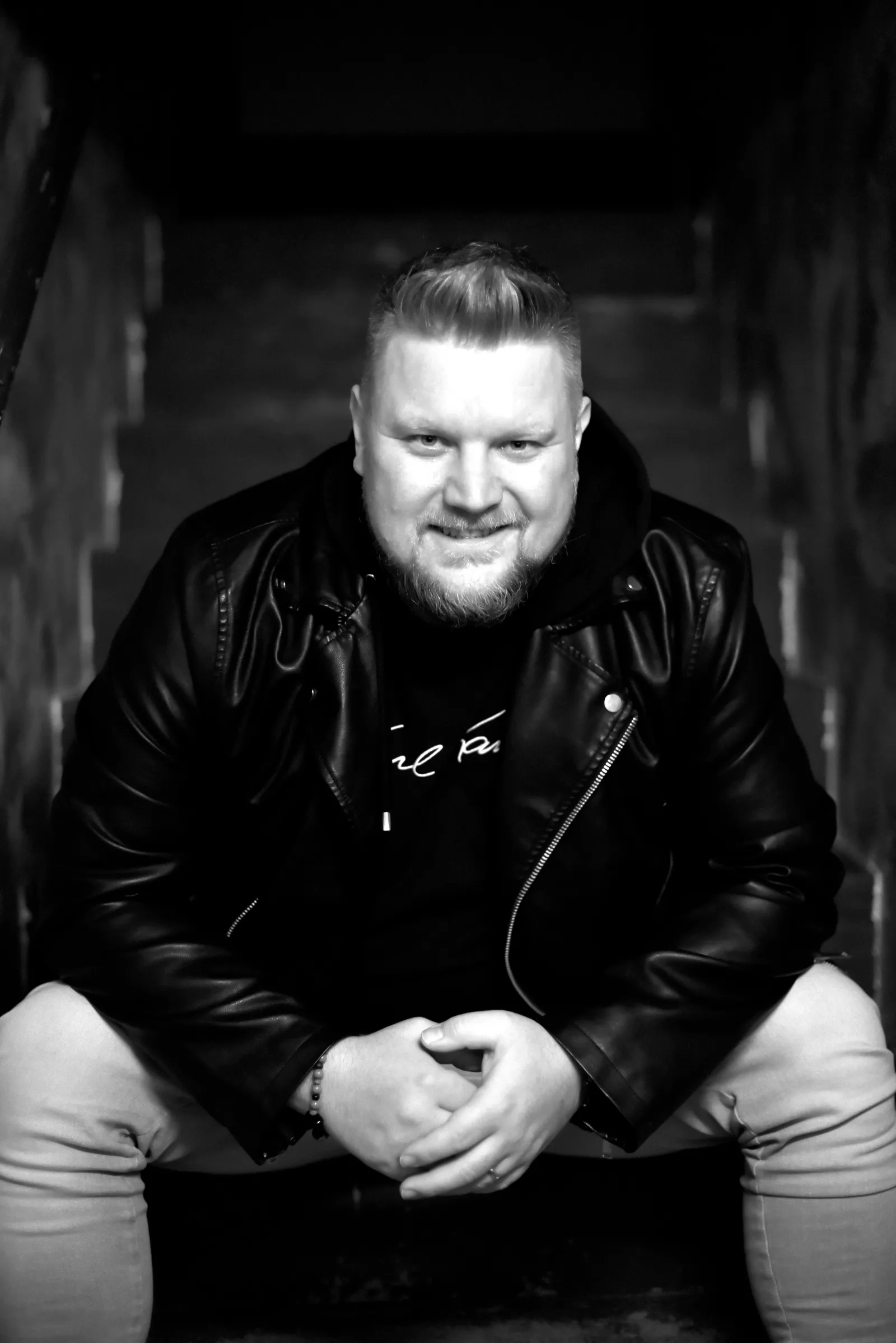 DJ Smatzo in Lederjacke sitzt auf einer Treppe, die Hände gefaltet, Schwarz-Weiß-Aufnahme.
