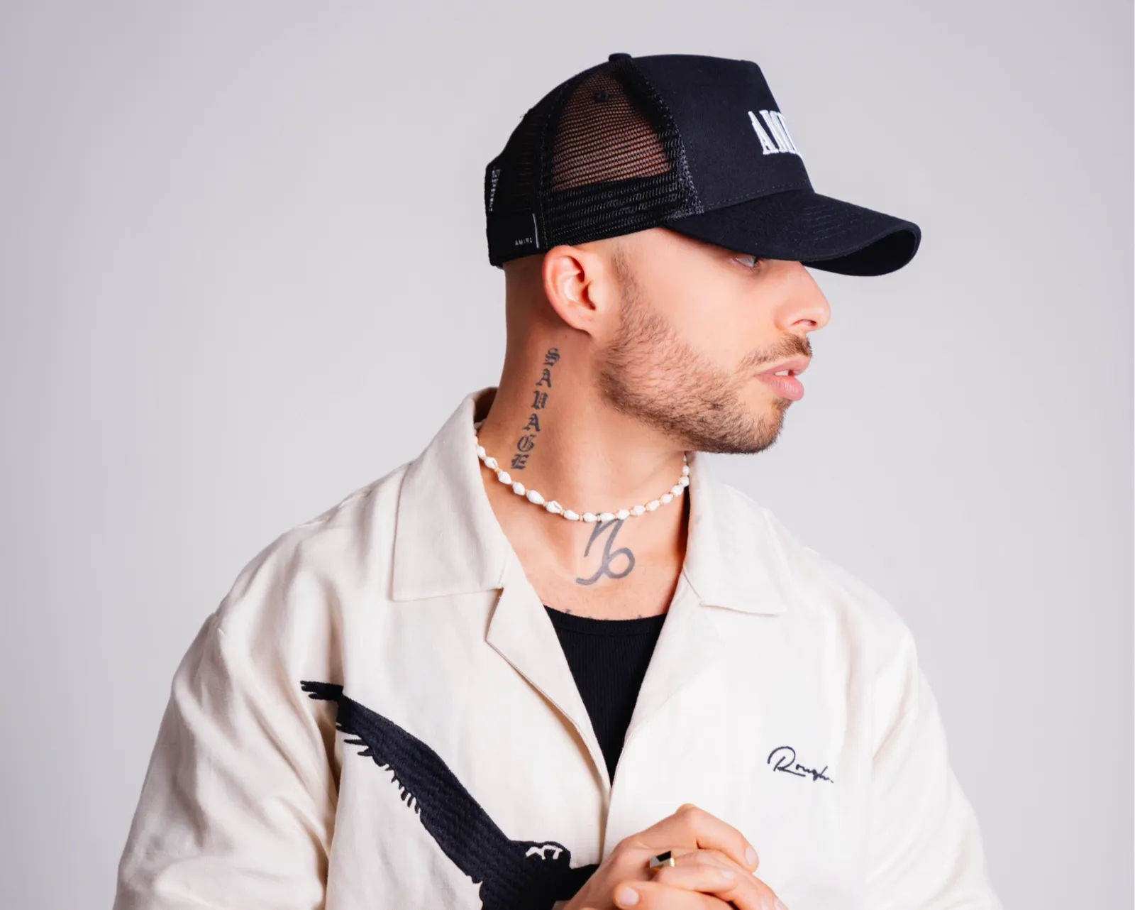 Mann mit schwarzer Cap im Profil, cremefarbenes Hemd, Perlenkette und Tattoo am Hals.