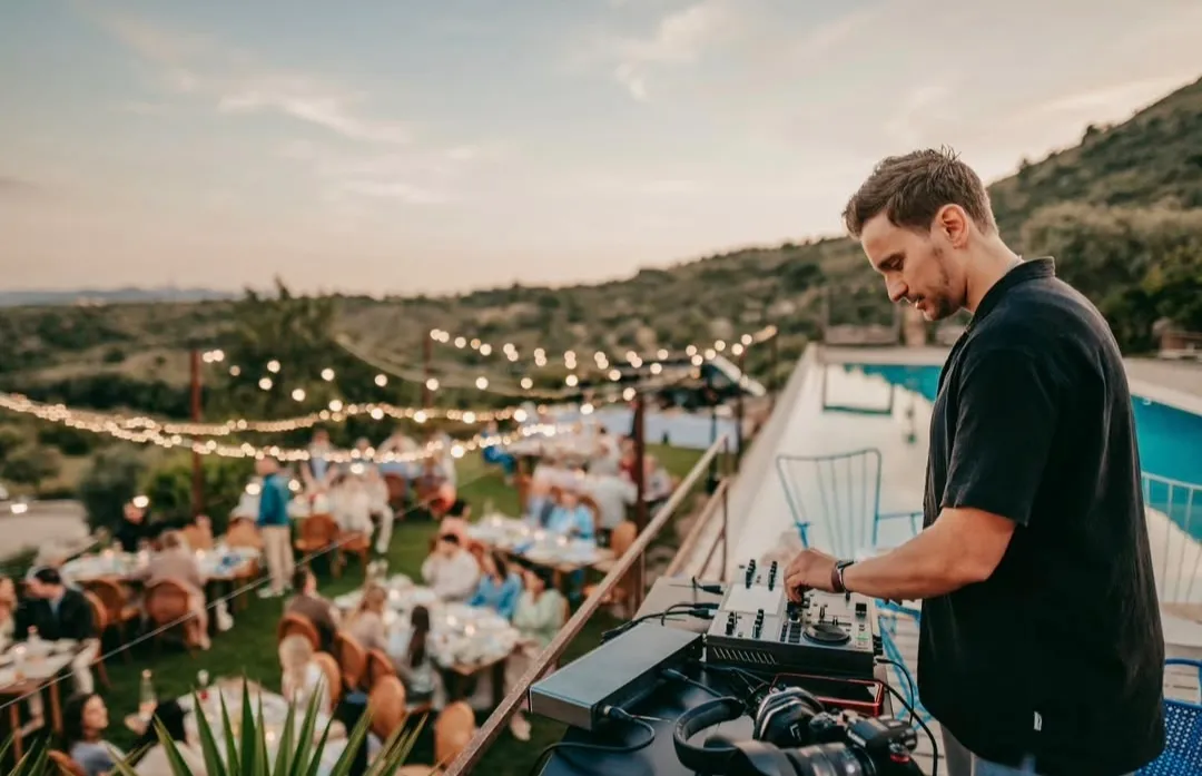 DJ Robinho am Controller auf einer Terrasse, darunter Gäste an Tischen unter Lichterketten.