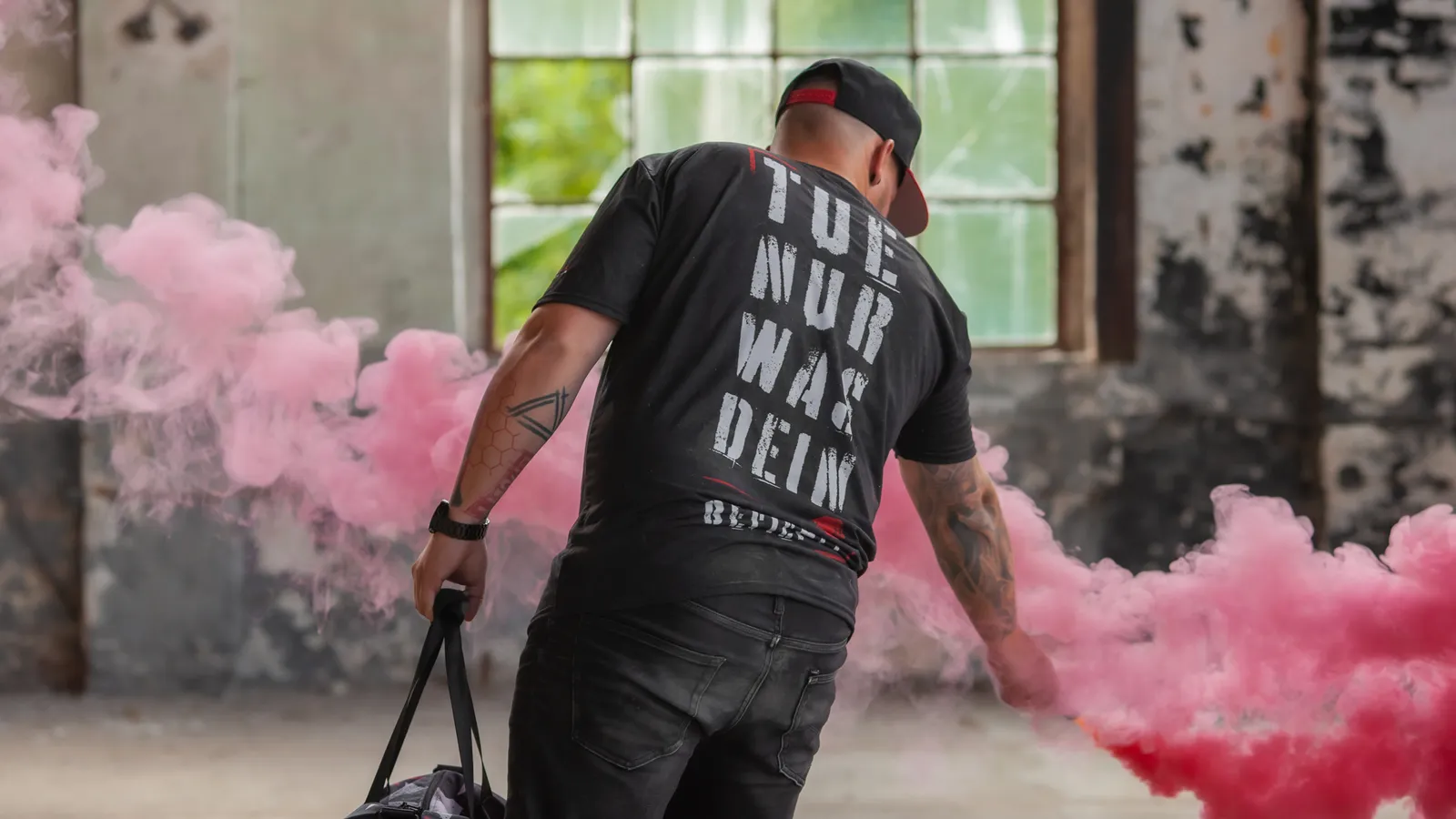 Mann von hinten in einer Industriehalle, in der Hand eine pinke Rauchfackel und eine Sporttasche.