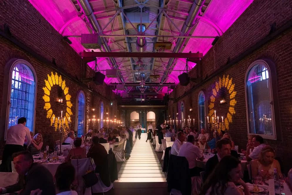 Backsteinhalle in Magenta beleuchtet, lange gedeckte Tafeln mit Gästen und weißer Mittelgang.
