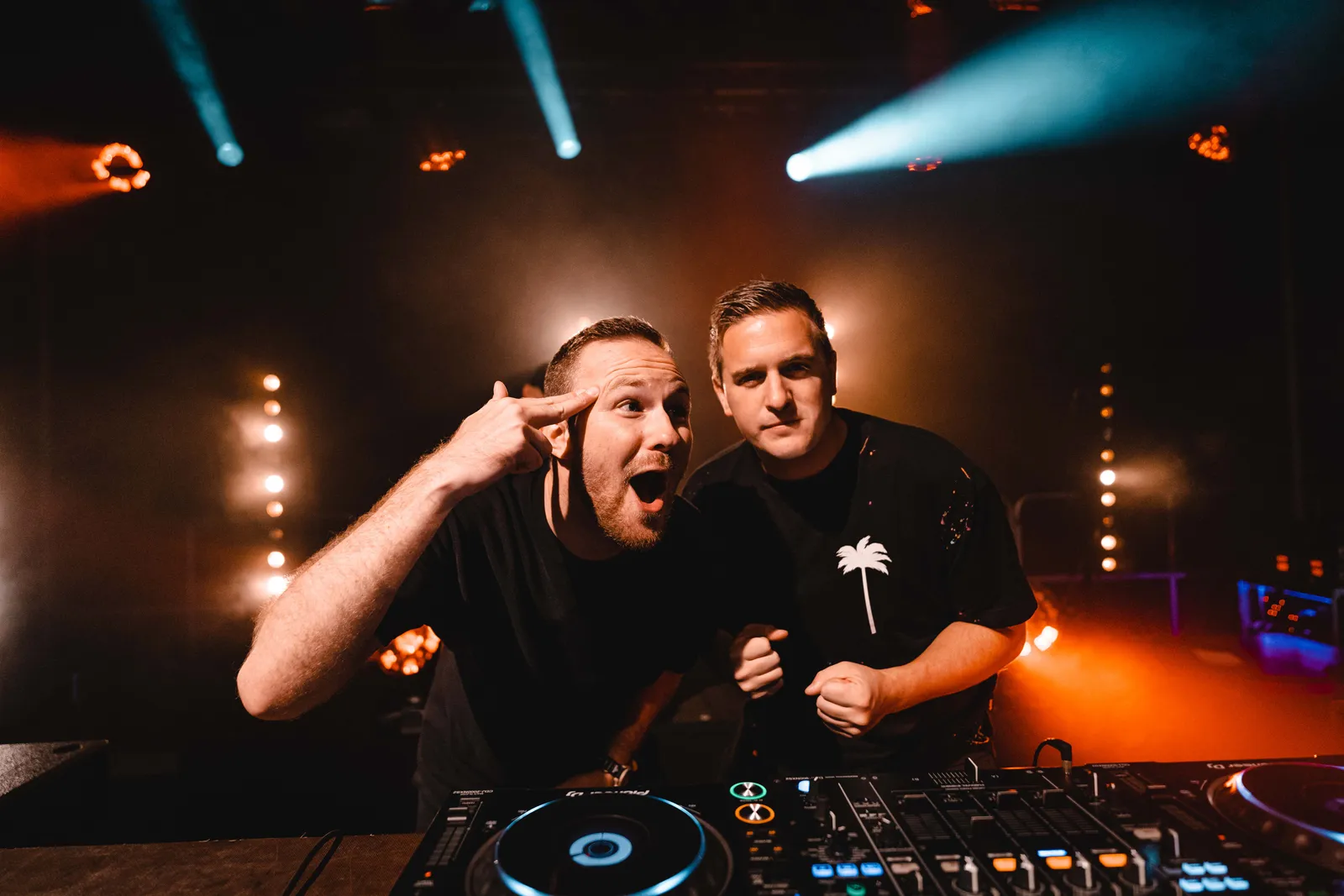 Zwei DJs am Pult in blau-orangem Clublicht, einer posiert mit Finger an der Schläfe.