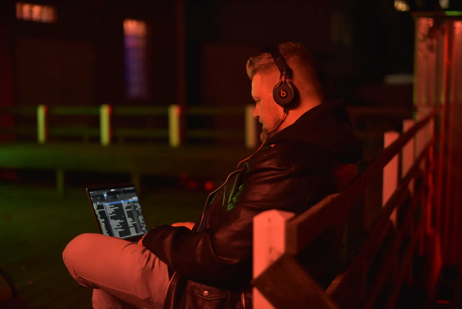 DJ Smatzo mit Kopfhörern auf einer Bank, Laptop mit Musiksoftware auf dem Schoß, rotes Licht.