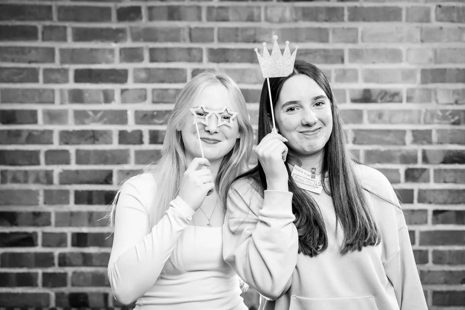 Zwei junge Frauen vor einer Ziegelwand halten Sternenbrille und Glitzerkrone an Stäben hoch, Schwarz-Weiß-Aufnahme.