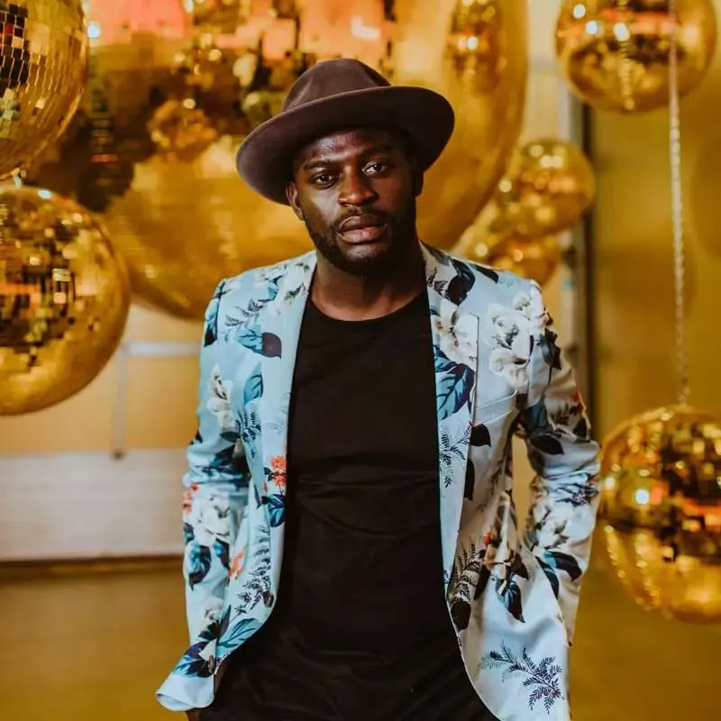 Mann mit braunem Hut und geblümtem Sakko vor einer Wand aus goldenen Discokugeln.