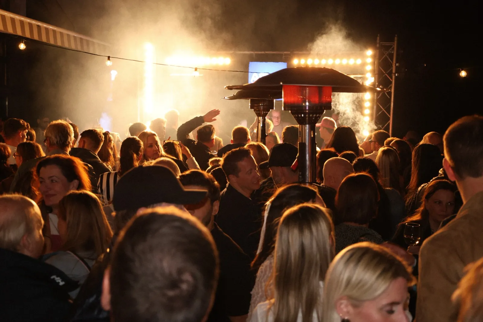 Dichtes Publikum bei einer Abendveranstaltung im Freien, Heizpilz, Lichterketten und Bühnenlicht.