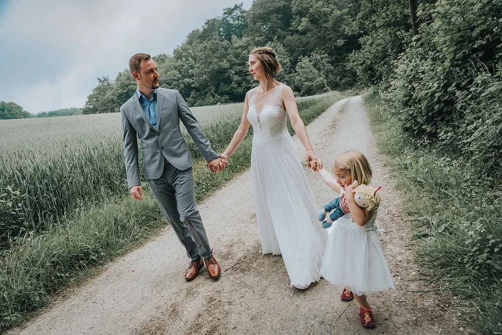 Brautpaar geht Hand in Hand einen Feldweg entlang, daneben ein Kind mit Puppe im Arm.