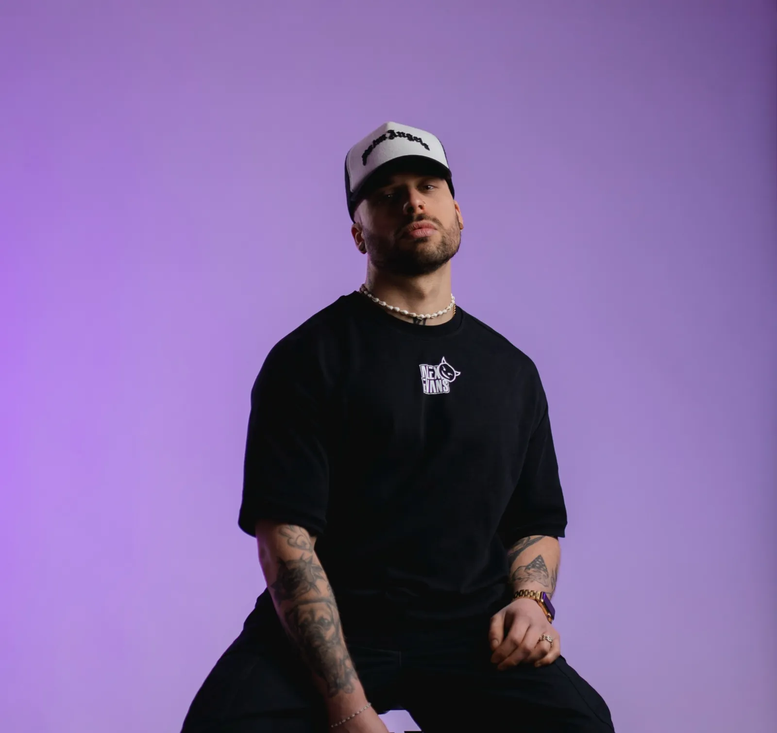 Alex Evans mit Cap und schwarzem Oversize-Shirt vor violettem Studiohintergrund.