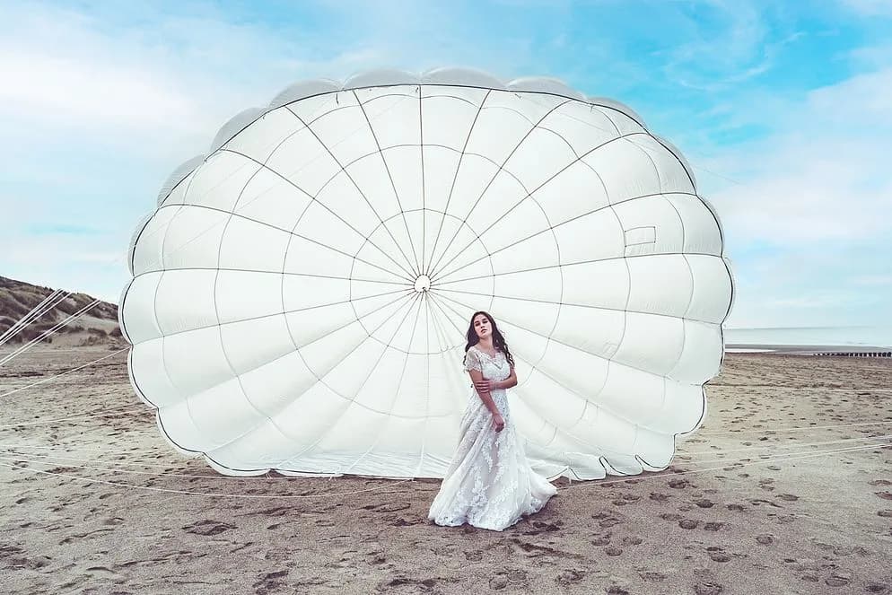 Braut im Spitzenkleid vor einem großen weißen Fallschirm am Sandstrand.