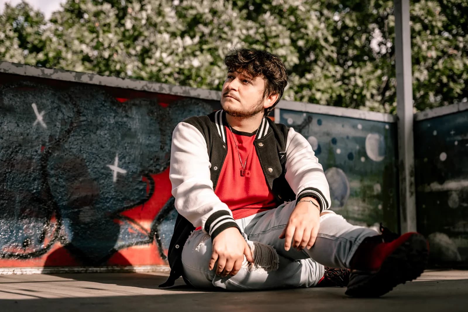 Junger Mann in College-Jacke sitzt im Schneidersitz vor einer Graffitiwand in der Sonne.