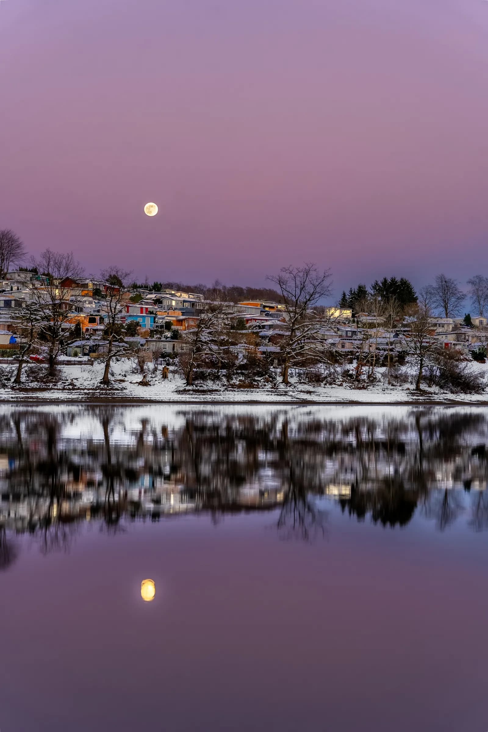 Vollmond am violetten Himmel über verschneiten Häusern am Hang, gespiegelt im ruhigen See.