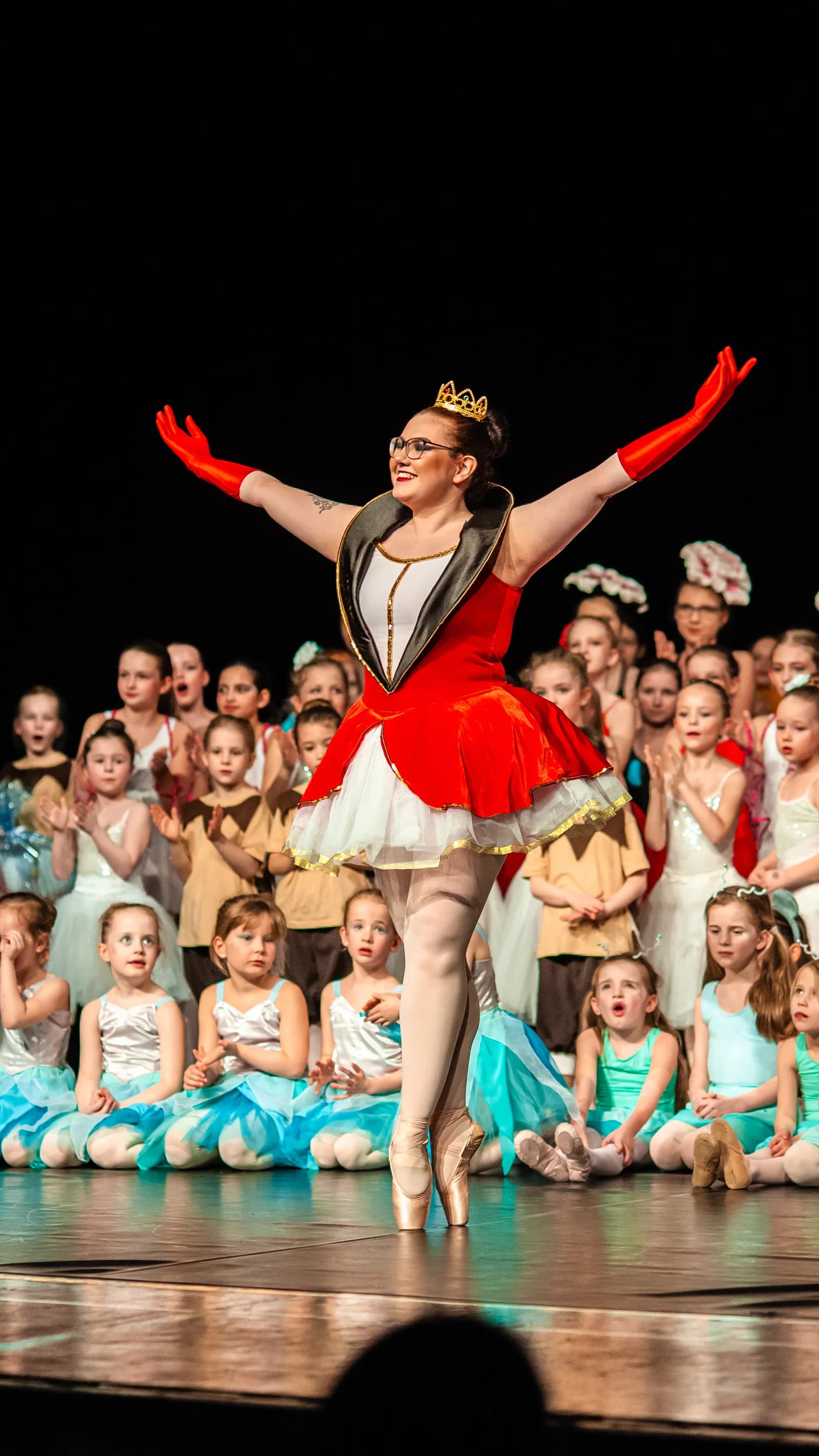 Tänzerin in rotem Tutu mit Krone steht auf Spitze, dahinter applaudierende Kinder in Kostümen.