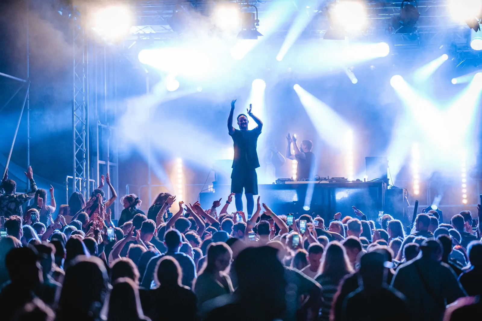 Mann mit erhobenen Händen auf einem Podest vor der Bühne, dahinter der DJ am Pult im Scheinwerferlicht.