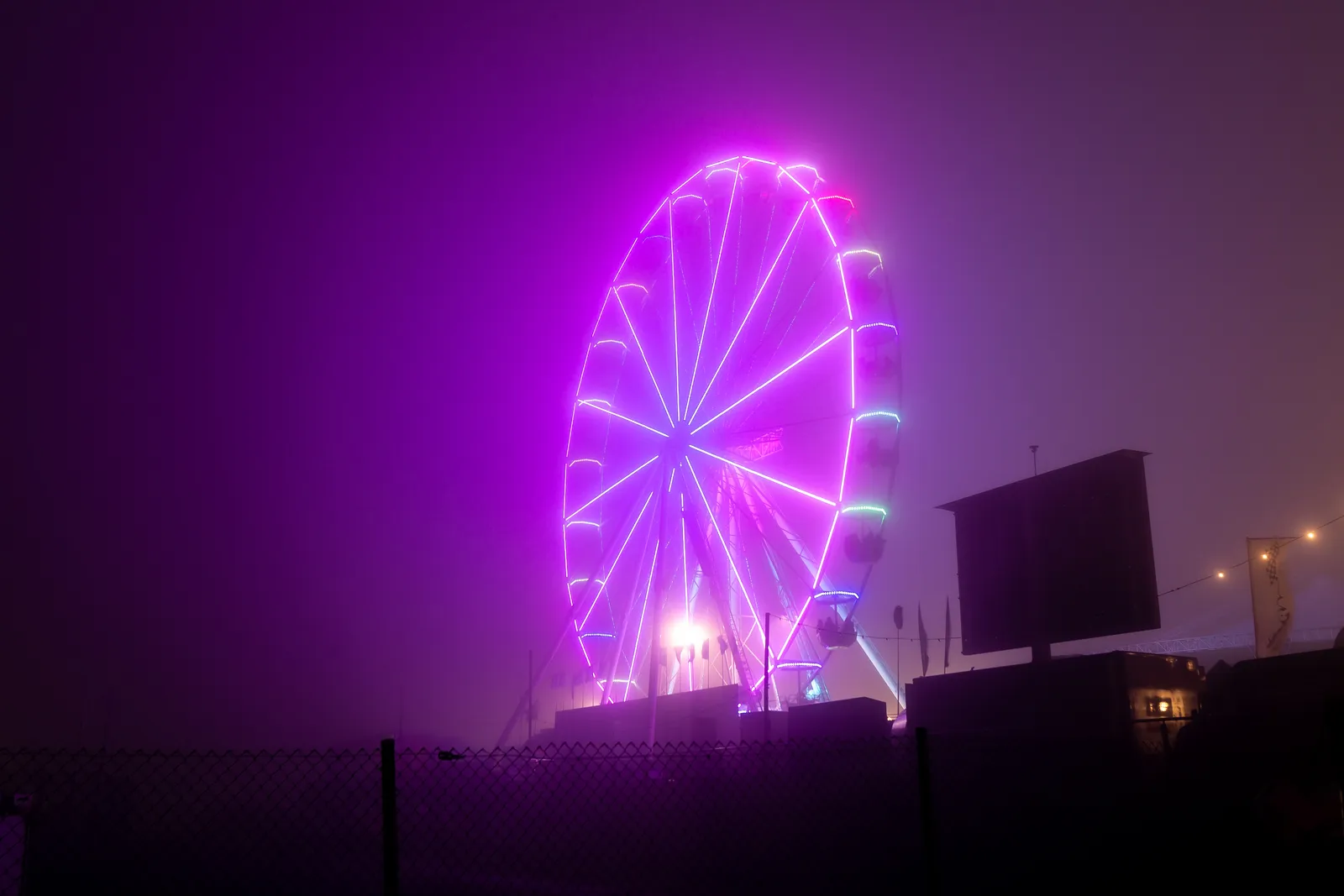 Beleuchtetes Riesenrad in Pink und Violett, nachts im Nebel hinter einem Maschendrahtzaun.