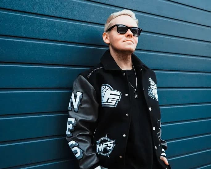 Tim Twice mit Sonnenbrille und schwarzer College-Jacke mit NFS-Emblemen vor blauer Wellblechwand.