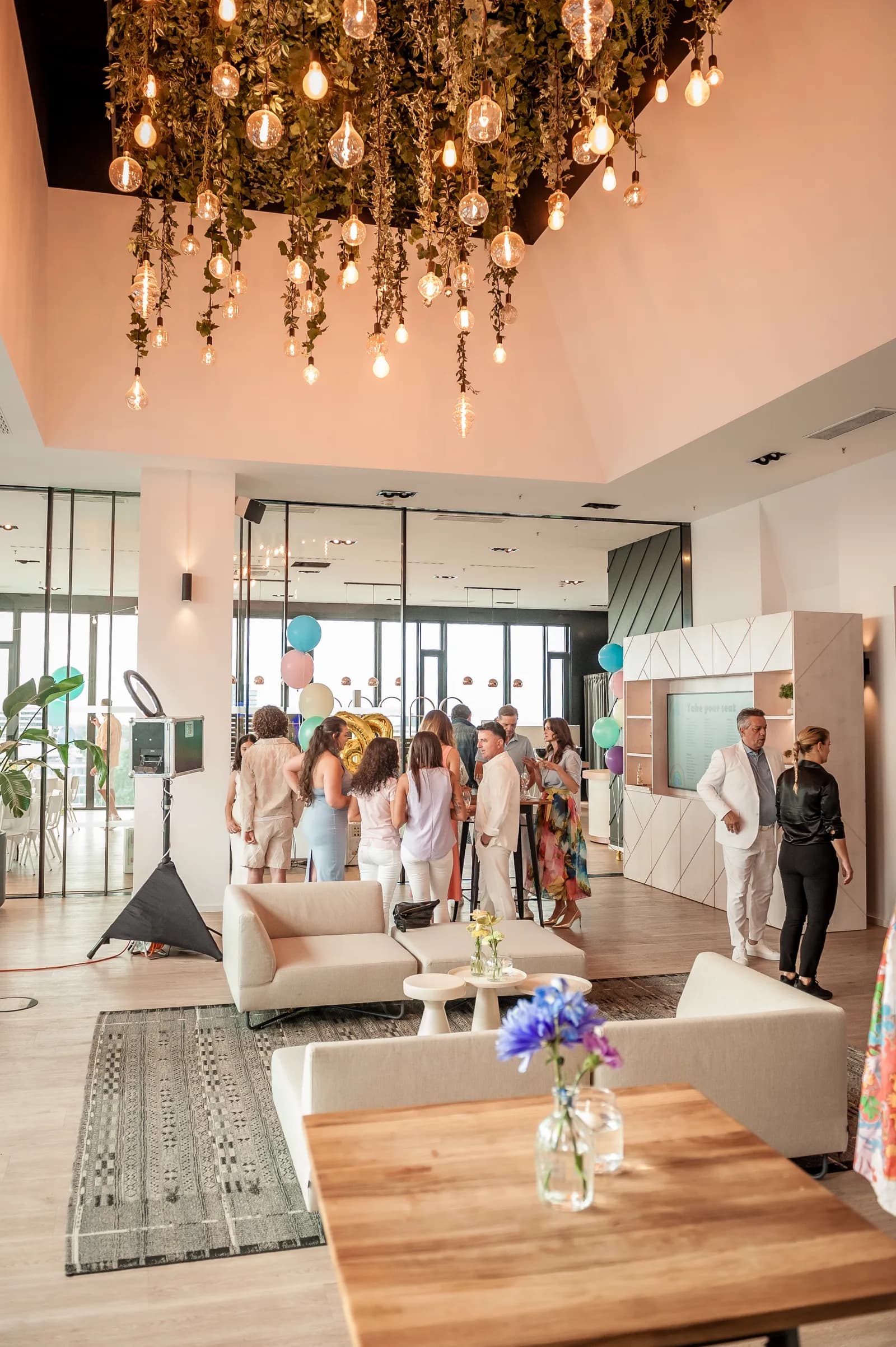 Heller Eventraum mit Glühbirnen-Deckeninstallation, Gäste zwischen hellen Sofas und Pastellballons.