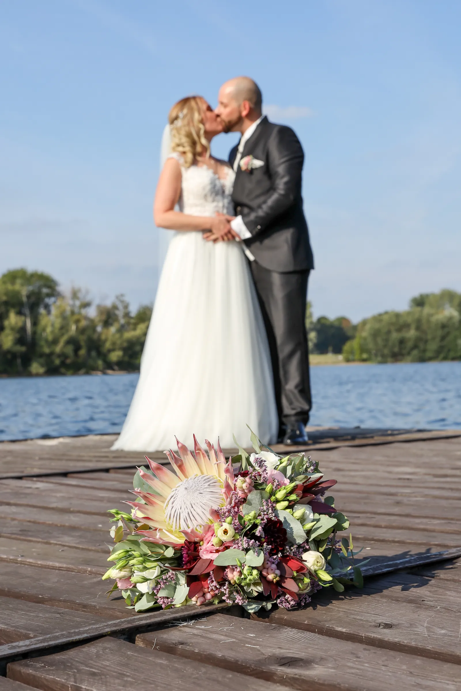 VIU Fotostudio: Brautstrauß mit Protea auf einem Holzsteg, dahinter ein küssendes Brautpaar am See.