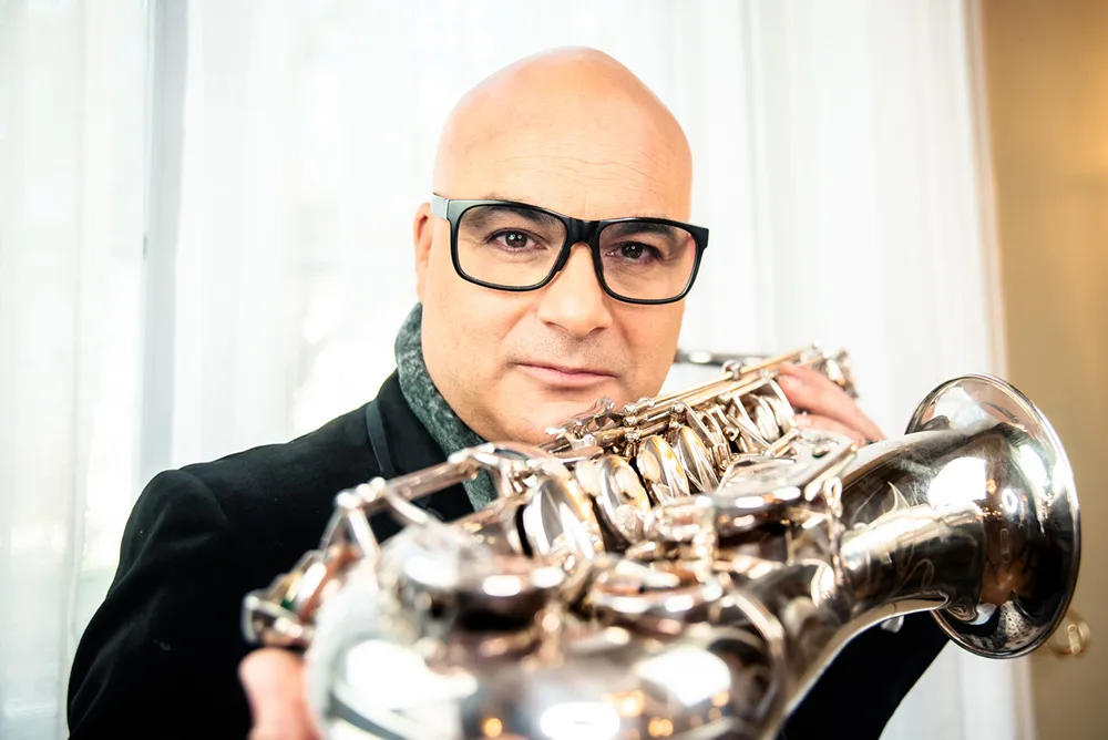 Gianni Bello mit schwarzer Brille und silbernem Saxophon vor einem hellen Vorhang.