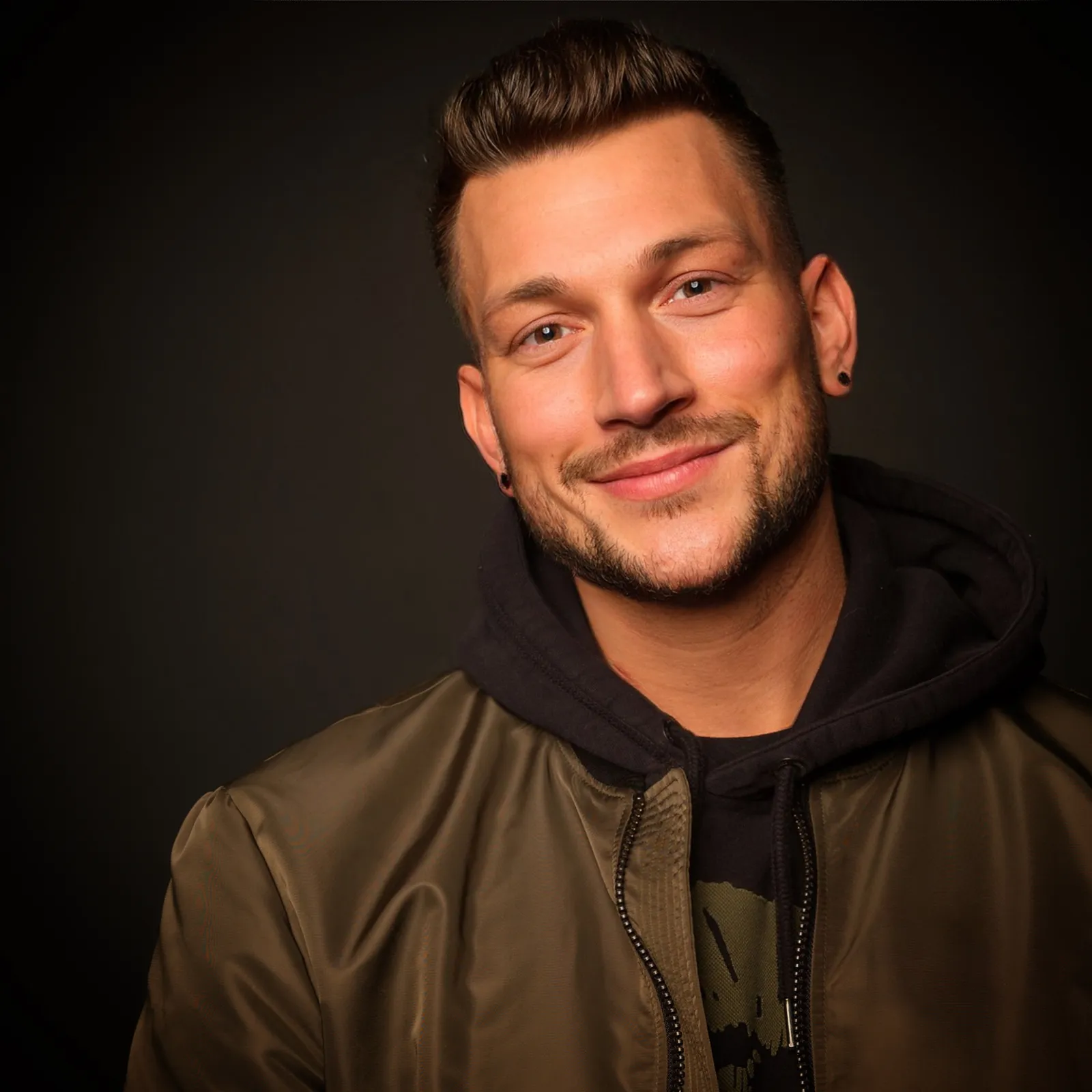 DJ Carl Bangz im Porträt, olivgrüne Bomberjacke über schwarzem Hoodie, dunkler Hintergrund.