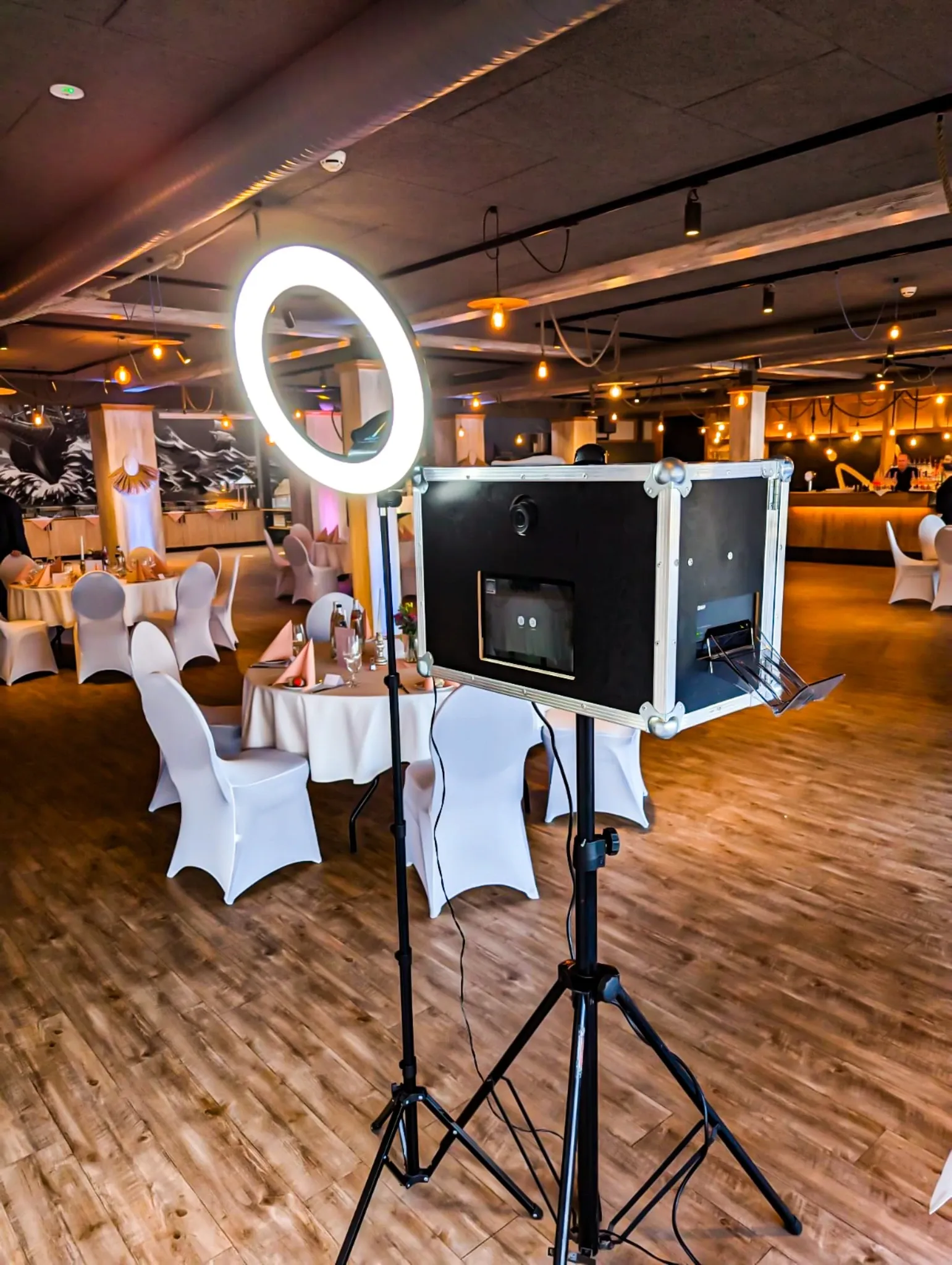 Fotobox mit Ringlicht auf Stativen in einem Saal mit eingedeckten Tischen und weiß bezogenen Stühlen.