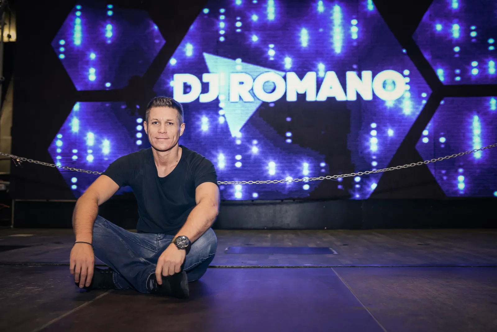 DJ Romano sitzt im Schneidersitz auf der Bühne vor einer LED-Wand mit seinem Schriftzug.