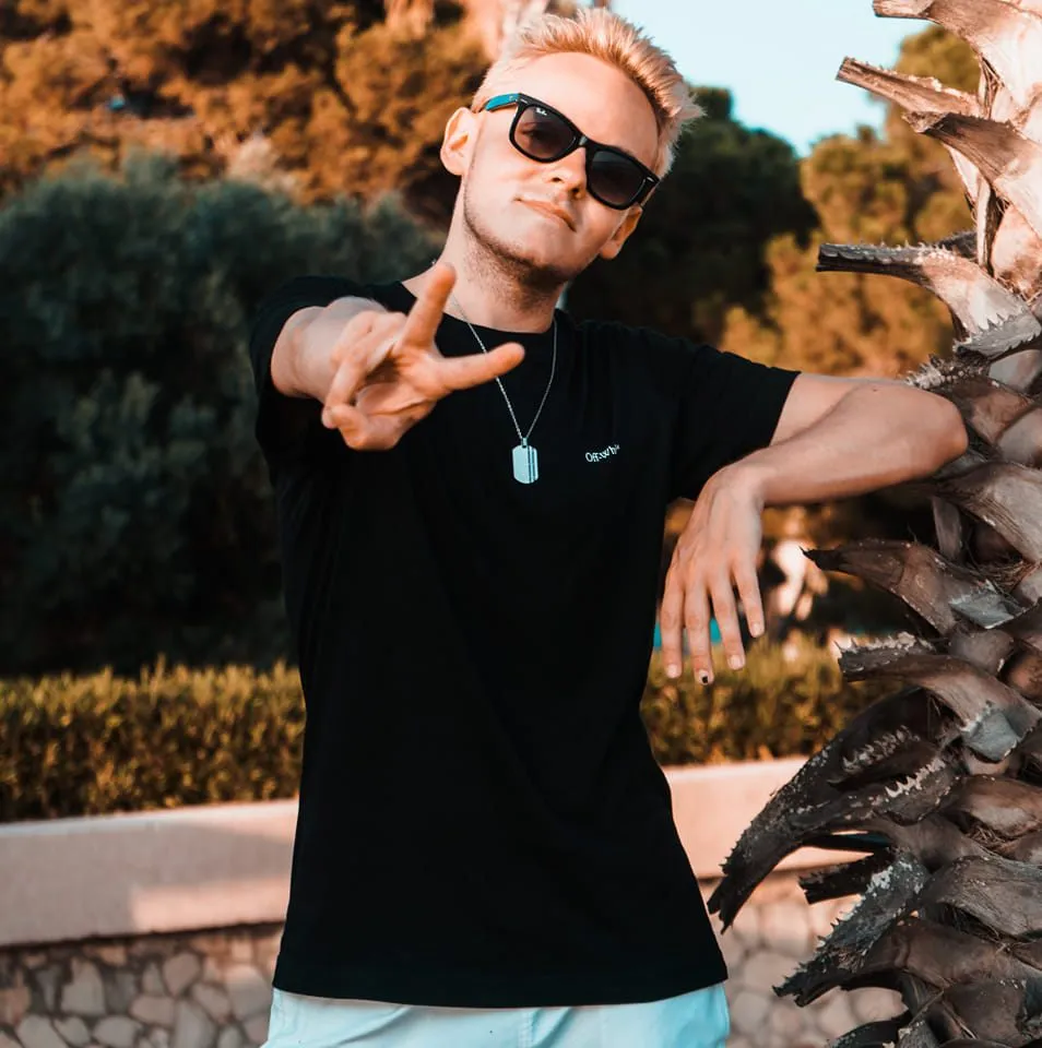Tim Twice mit blondem Haar und Sonnenbrille im schwarzen Shirt, Hand zur Kamera gestreckt, neben einer Palme.