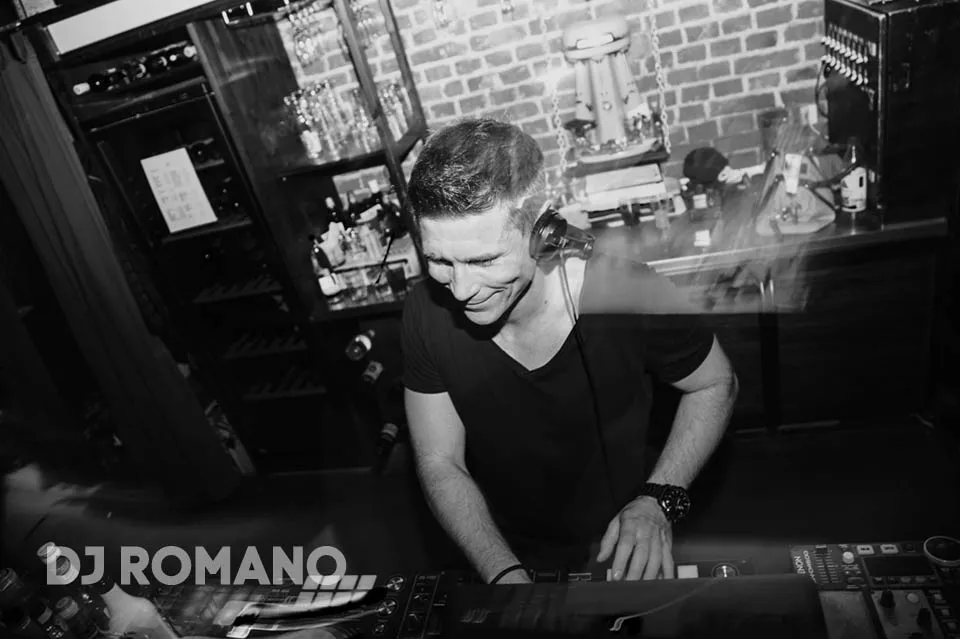 DJ Romano mit Kopfhörer am Pult vor einer Bar, Schwarz-Weiß-Aufnahme mit dem Schriftzug DJ Romano.