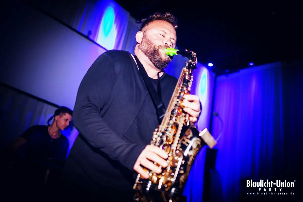 DimiSaxx spielt Saxophon im blauen Bühnenlicht, dahinter ein DJ am Pult.