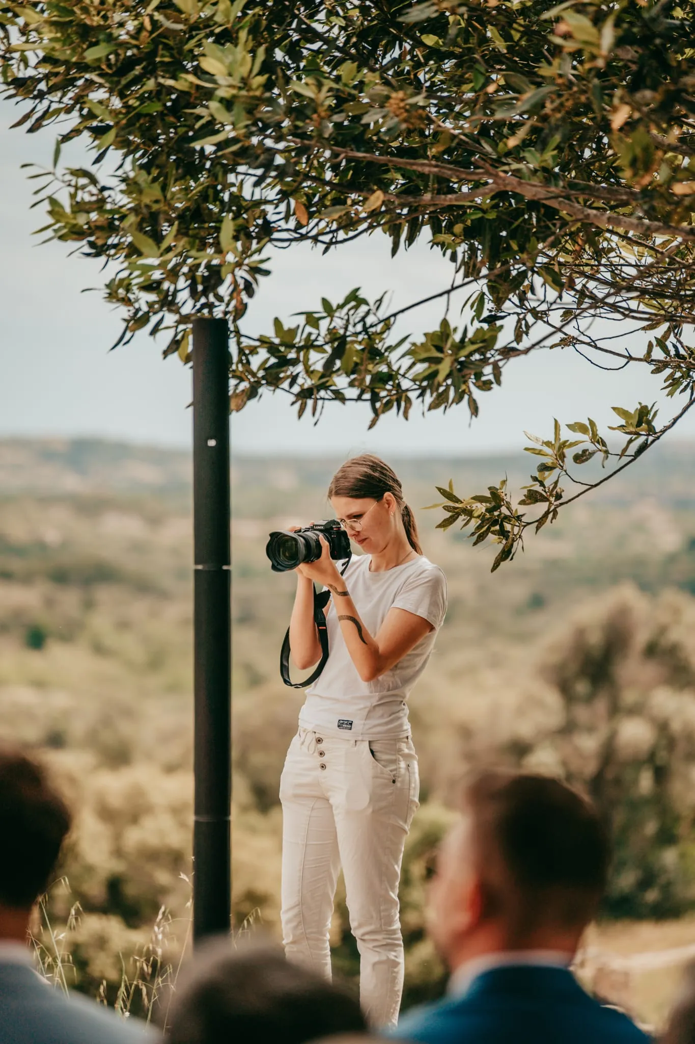 Ninanii Productions: Fotografin mit Kamera am Auge unter einem Baum, davor unscharfe Zuschauer.