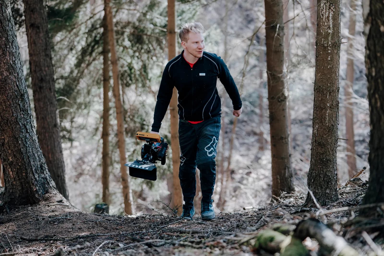 m.w.media.arts: Kameramann im Nadelwald, in der Hand eine Kamera mit Mikrofon, Monitor und Streulichtblende.