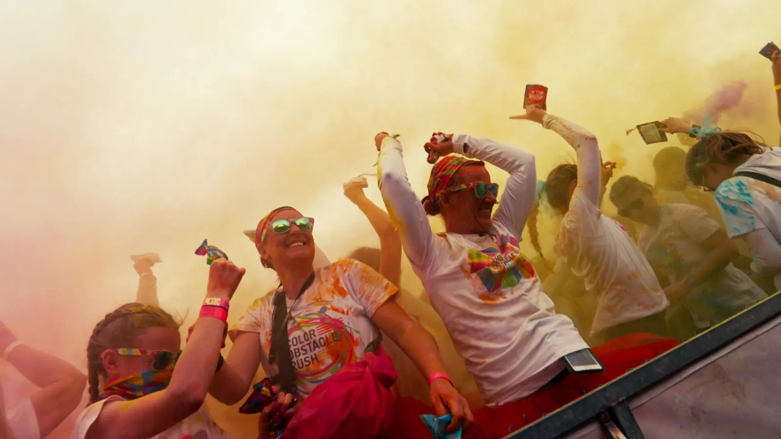 Läufer in Shirts mit Aufdruck Color Obstacle Rush werfen gelbes Farbpulver in die Luft.