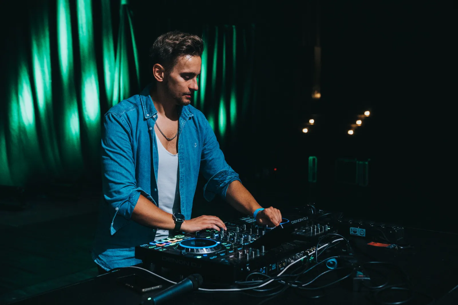 DJ Robinho am Denon-Controller, Jeanshemd über weißem Shirt, grün beleuchteter Vorhang.