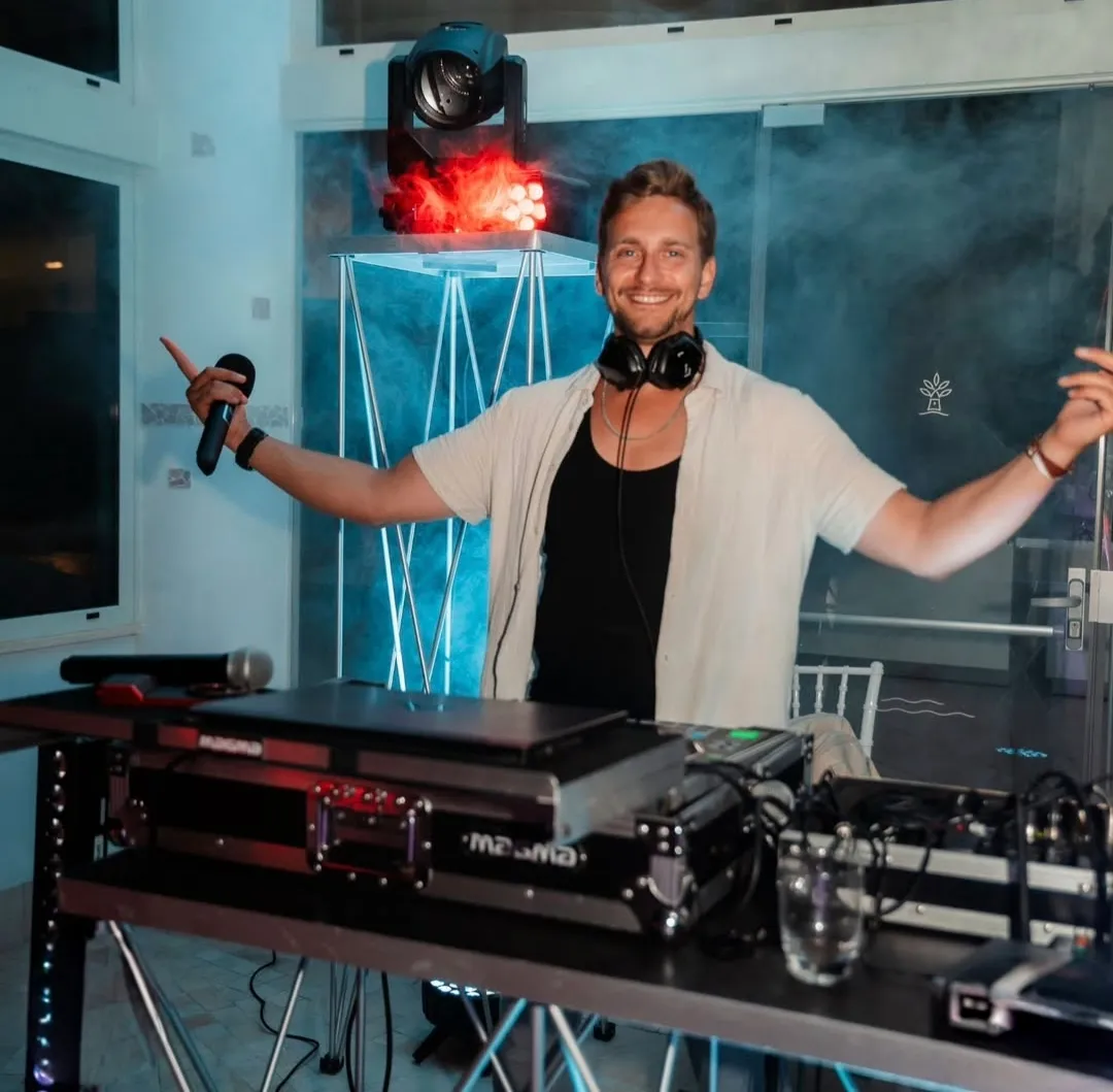 DJ Robinho hinter dem Pult, Mikrofon in der Hand, Arme ausgebreitet, Nebel und rotes Licht.