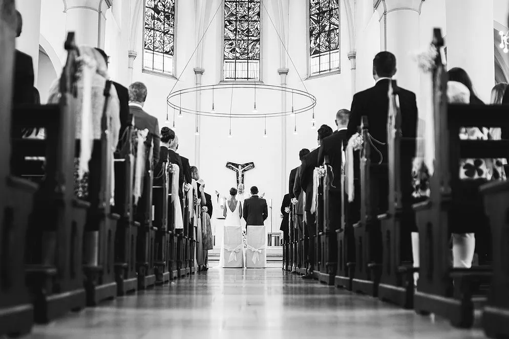 Pic-Factory: Blick durch den Mittelgang einer Kirche auf ein Brautpaar vor dem Altar, Gäste in den Bänken.