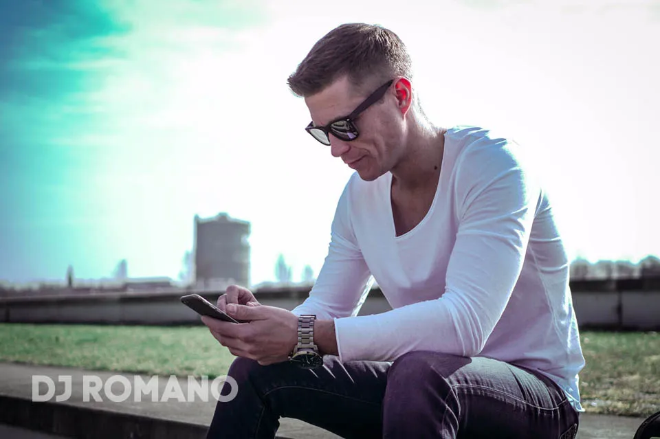 Mann mit Sonnenbrille im weißen Shirt sitzt im Freien am Smartphone, Schriftzug DJ Romano.