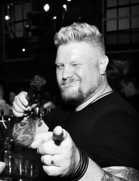 DJ Smatzo streckt ein Mikrofon in die Kamera und zeigt mit der anderen Hand nach vorn.