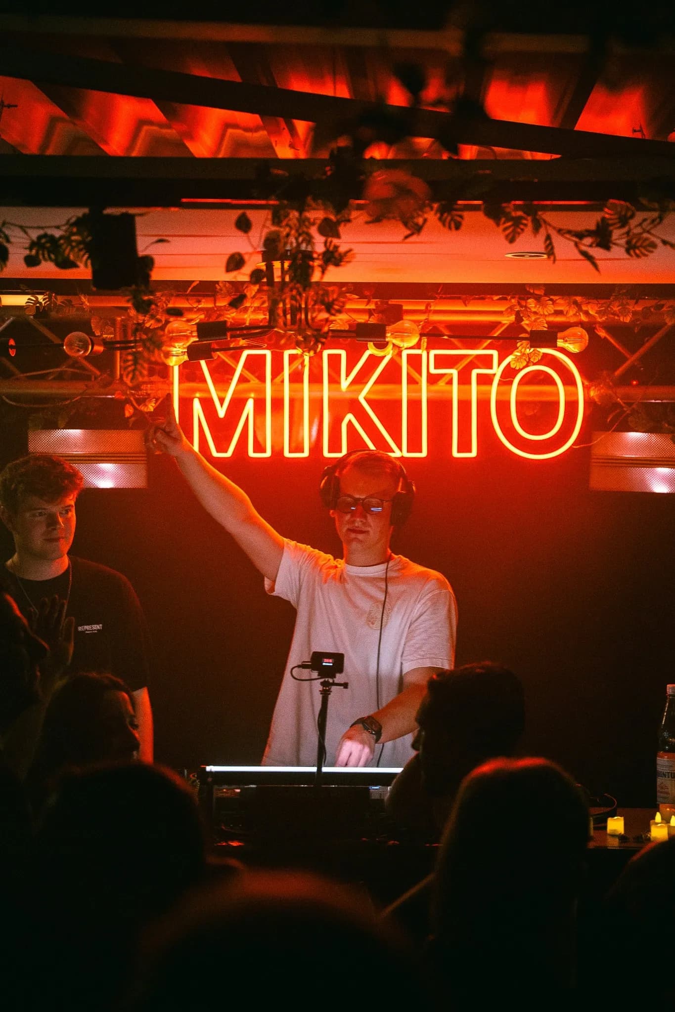 DJ am Pult hebt den Arm, dahinter leuchtet der rote Neonschriftzug MIKITO, davor Publikum.