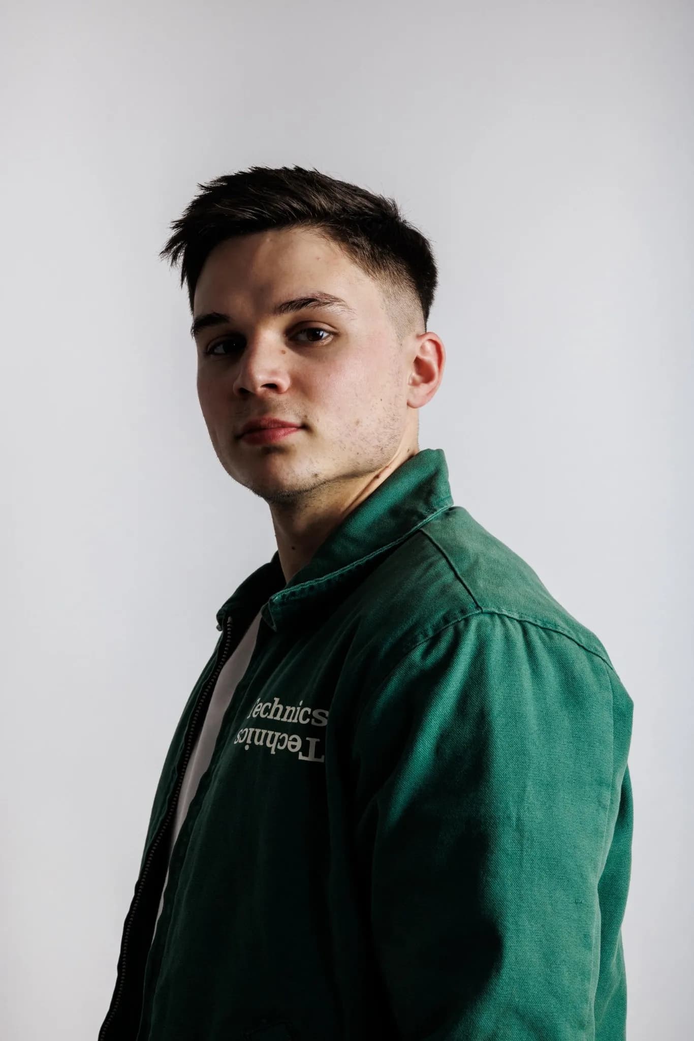 Junger Mann im Halbprofil in grüner Jacke mit Technics-Schriftzug vor heller Wand.