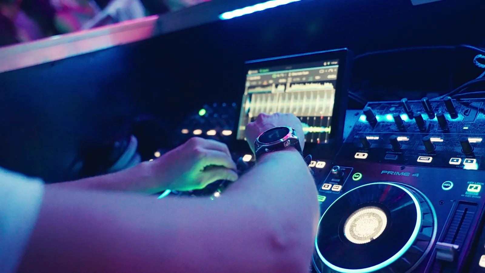 Hände eines DJs am Controller Prime 4, daneben ein Laptop mit Wellenform im blauvioletten Licht.